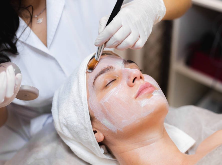 Beautician with a brush applies a white moisturizing mask to the face of a young girl client in a spa beauty salon